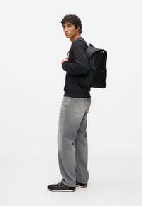 Czarny plecak o usztywnionej konstrukcji, matowym wykończeniu i srebrnych zamkach, noszony przez osobę ubraną w czarną bluzę i szare dżinsy.