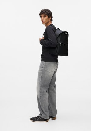 Fekete hátizsák, strukturált dizájnnal, matt felülettel és ezüst cipzár részletekkel, egy fekete pulóvert és szürke farmernadrágot viselő személynél.