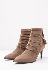 Botas de tobillo de ante en marrón claro, con punta afilada, diseño envuelto con cordones entrelazados y un borlón decorativo. Tacón alto y delgado.