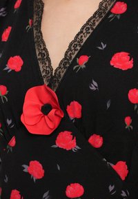 Černé květinové šaty s červenými růžovými vzory, krajkovým lemováním kolem výstřihu a výrazným červeným květinovým akcentem na prsou.