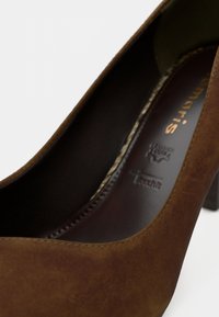 Chaussure à talon haut en daim marron avec un bout arrondi et un intérieur brillant marron foncé. Présente un logo estampé sur la semelle intérieure.