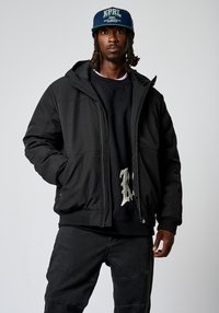 Chaqueta de plumas negra con un acabado texturizado, cierre de cremallera y capucha. Llevada sobre un sudadera negra con un diseño ligero. Pantalones oscuros a juego.