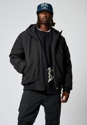 Zwarte pufferjack met een gestructureerde afwerking, ritsluiting en capuchon. Draag het over een zwarte sweatshirt met een licht ontwerp. Bijpassende donkere broek.