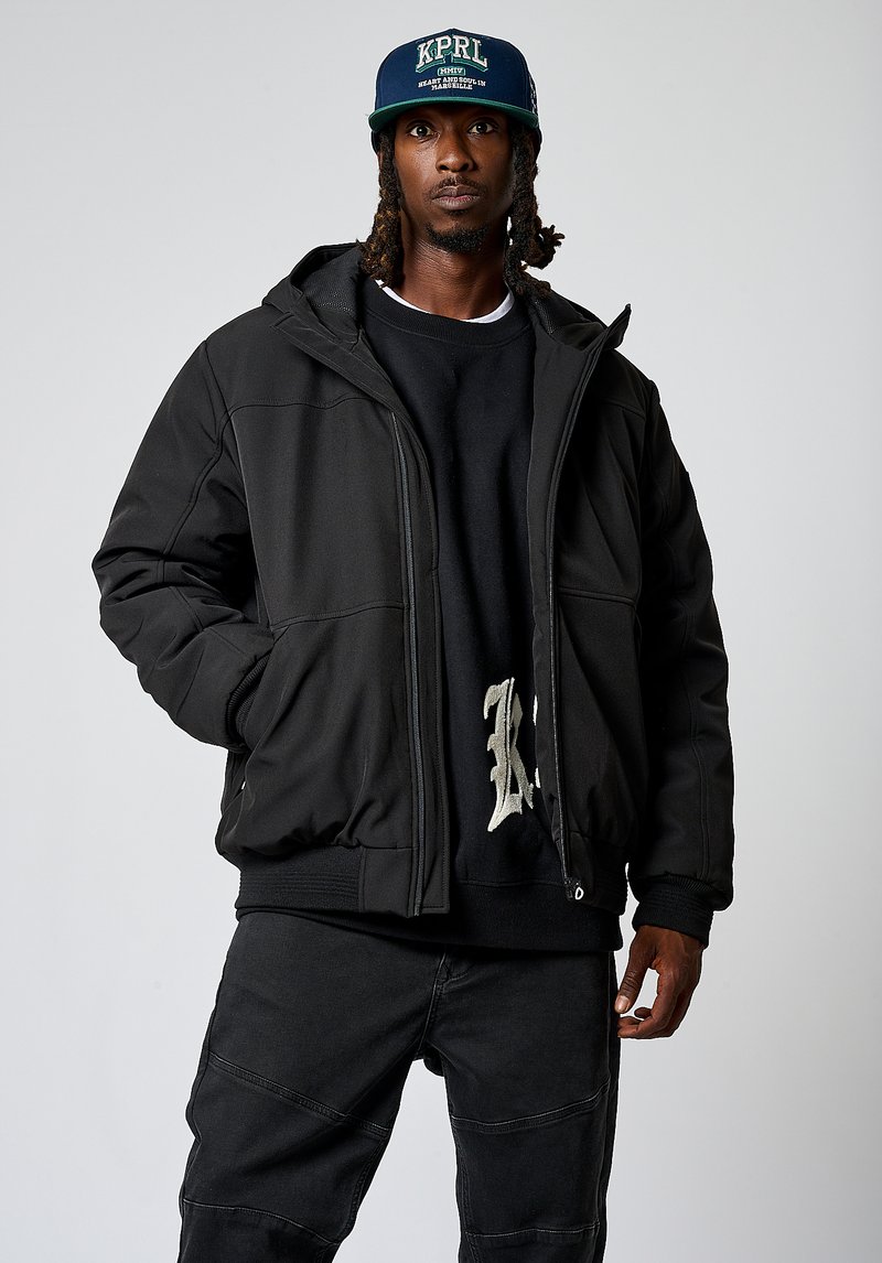 Chaqueta de plumas negra con un acabado texturizado, cierre de cremallera y capucha. Llevada sobre un sudadera negra con un diseño ligero. Pantalones oscuros a juego.