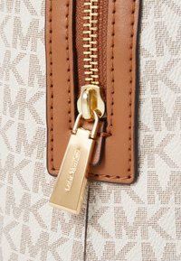 Matériau texturé blanc avec un motif géométrique, accent en cuir marron près d'une tirette dorée avec un détail de marque gravé.