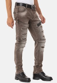 Ausgebleichte, braune, ausgewaschene Jeans mit Rissen, detaillierten Nähten und einer Seitentasche. Kombiniert mit einem schwarzen, strukturierten Gürtel und schwarzen Schuhen.