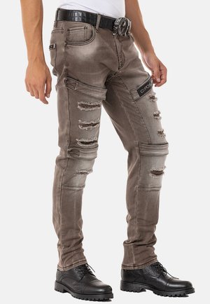 Ausgebleichte, braune, ausgewaschene Jeans mit Rissen, detaillierten Nähten und einer Seitentasche. Kombiniert mit einem schwarzen, strukturierten Gürtel und schwarzen Schuhen.