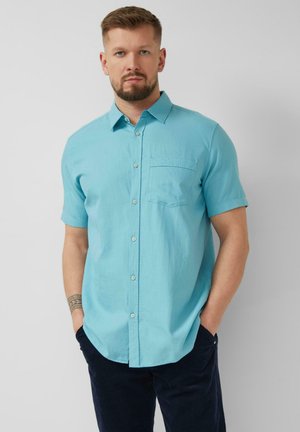 Mann mit Bart, trägt ein hellblaues kurzärmliges Hemd mit Knöpfen und dunkle Hosen, steht mit den Händen in den Taschen vor einem einfarbigen Hintergrund.