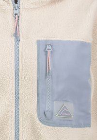 Beige Fleecejacke mit einer hellblauen Brusttasche mit Reißverschluss, die rosa Reißverschlussanhänger und ein dreieckiges "Outdoor"-Logoabzeichen aufweist.