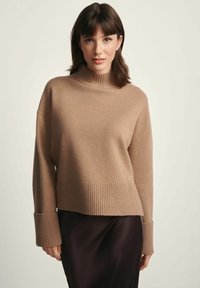 Młoda kobieta z brązowymi włosami nosi beżowy sweter typu golf z prążkami oraz ciemną spódnicę, stojąc na tle jasnego, jednolitego tła.
