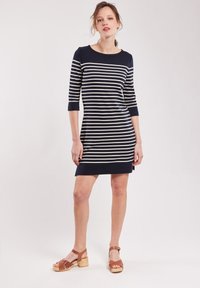 Robe rayée bleu marine et crème avec des manches trois-quarts, décolleté rond, longueur genoux, assortie à des sandales à talons marron.