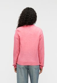 Ροζ μάλλινο πουλόβερ με υφή, με στρογγυλή λαιμόκοψη και ριμπ μανσέτες. Φορώντας το πάνω από  ανοιχτόχρωμο τζιν, εμφανίζεται από πίσω.