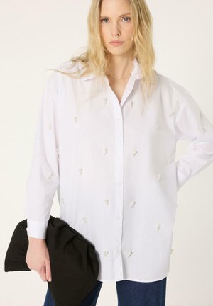 Femme blonde portant une chemise blanche oversize avec de petits regroupements de perles, tenant une grande pochette noire, debout devant un fond uni.