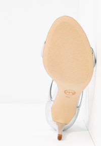 Semelle d'une chaussure à talon haut argentée soulevée, montrant la semelle beige texturée et le petit embout du talon, taille 7M indiquée près de la zone de la voûte plantaire.