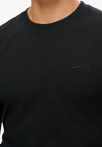 Närbild av en person som bär en svart t-shirt med rund hals och Superdry-märke, med en broderad logo på bröstet.