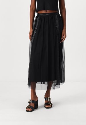 Frau trägt ein schwarzes Cropped-Top, schwarzen durchsichtigen Midirock und schwarze Sandalen mit Absatz und goldenen Schnallen, steht vor einem schlichten Hintergrund.