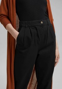 Pantalon noir sur mesure avec un devant plissé, doté d'une fermeture à un bouton et de poches latérales, associé à un cardigan couleur rouille.