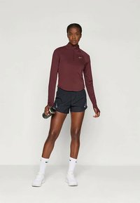 Egy nő bordó hosszúujjú felsőt, fekete sportnadrágot, fehér zoknit és fehér sportcipőt visel, miközben egy fekete vízpalackot tart a kezében, egy sima háttér előtt állva.