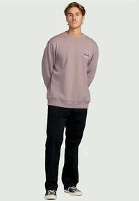 Jonge man die met zijn handen achter zijn rug staat, gekleed in een mauve sweatshirt, zwarte broek en zwart-witte sneakers, tegen een effen achtergrond.
