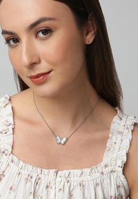 Silberne Schmetterling-Halskette mit detailliertem Design, bestehend aus zwei Flügeln und einem kleinen klaren Edelstein, an einer zarten Kette. Rundhalsausschnitt mit gerafftem Saum.