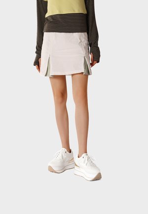 Donna con mini gonna bianca con pieghe laterali, top a maniche lunghe a righe e sneakers bianche con suola platform, in piedi contro uno sfondo semplice.