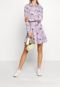 Robe violette à manches courtes avec un motif représentant des arbres et des figures. Assortie à un petit sac à main vert clair et des baskets blanches.