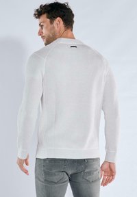 Weißer gerippter Pullover mit langen Ärmeln und rundem Ausschnitt, mit einem dezenten Etiketten-Detail auf der Rückseite. Kombiniert mit grauen, eng anliegenden Hosen.