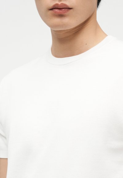 Κοντινό πλάνο ενός ατόμου που φοράει ένα απλό λευκό t-shirt με λαιμόκοψη, εμφανίζοντας το λαιμό, τη γραμμή του σαγονιού και τον άνω κορμό.