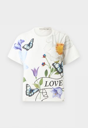 Žena nosi biele tričko s krátkym rukávom s motívmi motýľov a kvetín a textom "LOVE", v kombinácii s tmavými širokými džínsami a bielymi podpätkami.
