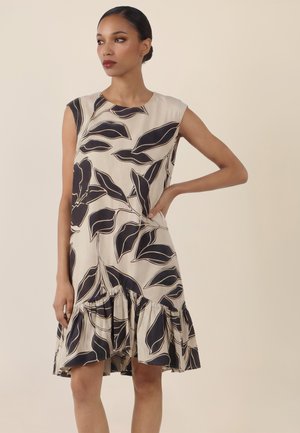 Frau steht mit der Hand in der Hüfte, trägt ein ärmelloses beiges Kleid mit großen schwarzen Blattmustern und einem gerafften Saum.