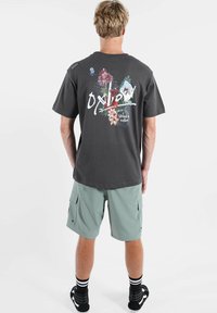 T-shirt en coton gris avec un motif floral et le texte "Oxbow" au dos, associé à un short cargo vert clair et des chaussettes noires à rayures blanches.