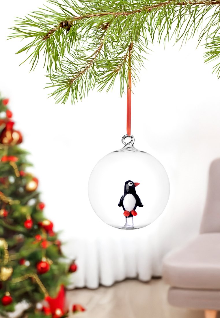 Klarer Glasornament mit einer Schwarz-Weiß-Pinguinfigur im Inneren. Oranger Schnabel und Füße. Hängt an einem roten Band von einem Kiefernzweig.