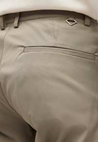 Κοντινή λήψη μπεζ παντελονιού που δείχνει την πίσω τσέπη με οριζόντια άνοιγμα και έναν μικρό ασημένιο τρίγωνο μεταλλικό κρίκο κοντά στη μέση.