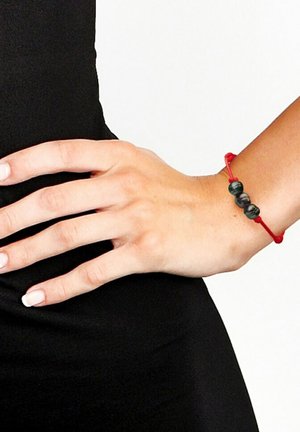 Bracelet élastique rouge avec trois perles sombres, texture lisse, design simple, porté sur une main reposant sur un fond noir.