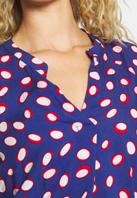 Blå blus med cirkulära mönster i röd och vit. Lätt V-halsdesign med synliga sömmar. Lättvikts tyg.