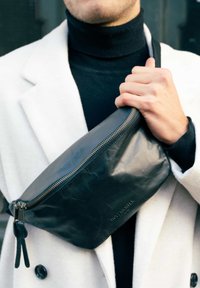 Schwarze Leder-Gürteltasche mit glatter Textur, goldfarbenem Reißverschluss und dezent geprägtem Markenlogo, quer über den Körper in einem lässigen Umfeld getragen.