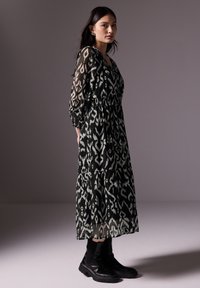 Lange jurk met zwart- en crèmekleurig patroon en een gelaagd ontwerp, diepe V-hals en doorschijnende mouwen, gecombineerd met zwarte enkellaarsjes.