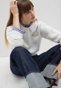 Weißer Pullover mit Kragen und goldenen Stickereien, über einem blau gestreiften Hemd getragen. Dunkelblaue Jeans mit hellblauen Umschlägen.