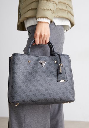 Personne portant une veste beige et un pantalon gris tenant un sac à main gris de la marque Guess avec des poignées supérieures et une étiquette logo.