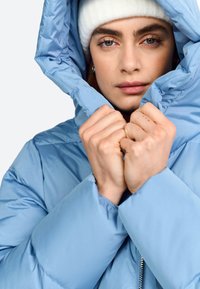 Veste padded bleu clair avec capuche, dotée d'une finition brillante et de poignets élastiques. Le modèle tient le col de la veste près de son visage.