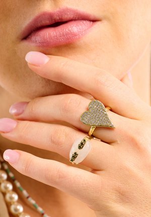 Gold rings featuring a heart-shaped design with small gemstones and an oval white stone adorned with green accents, worn on a hand.