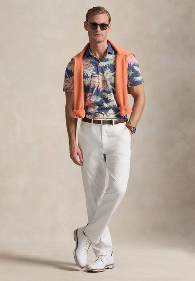 Άνδρας με φλοράλ κοντομάνικο πουκάμισο, λευκό παντελόνι, λευκά παπούτσια, γυαλιά ηλίου, πορτοκαλί πουλόβερ περασμένο πάνω από τους ώμους, στέκεται με το ένα χέρι στην τσέπη.