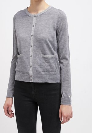 Kobieta ubrana w dopasowany szary sweter na guziki z dwiema przednimi kieszeniami oraz czarne spodnie na jednolitym tle.