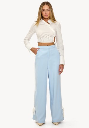 Blouse blanche à manches longues, courte et croisée, associée à un pantalon large bleu clair avec des rayures sur les côtés et des nœuds aux ourlets.