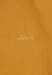 Μουσταρδί κίτρινο υφασμάτινο ύφασμα με μικρό, καλλιγραφικό κεντημένο κείμενο "Blkvis" σε ταιριαστό νήμα.