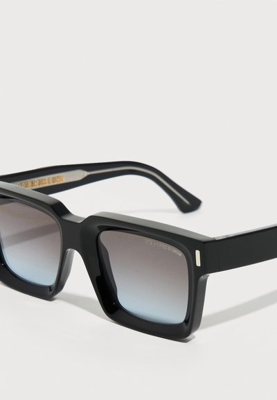 Μαύρα ορθογώνια γυαλιά ηλίου με παχιά πλαίσια και φακούς με γκράντιεντ γκρι, τοποθετημένα σε λευκή επιφάνεια.