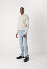 Suéter de cuello alto gris claro con puños acanalados, combinado con jeans rectos azul claro y zapatos de vestir negros, de pie sobre un fondo blanco.