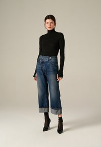 Donna in piedi che indossa un dolcevita nero, jeans a gamba larga blu con risvolto, calze nere trasparenti e décolleté neri a punta su uno sfondo beige.