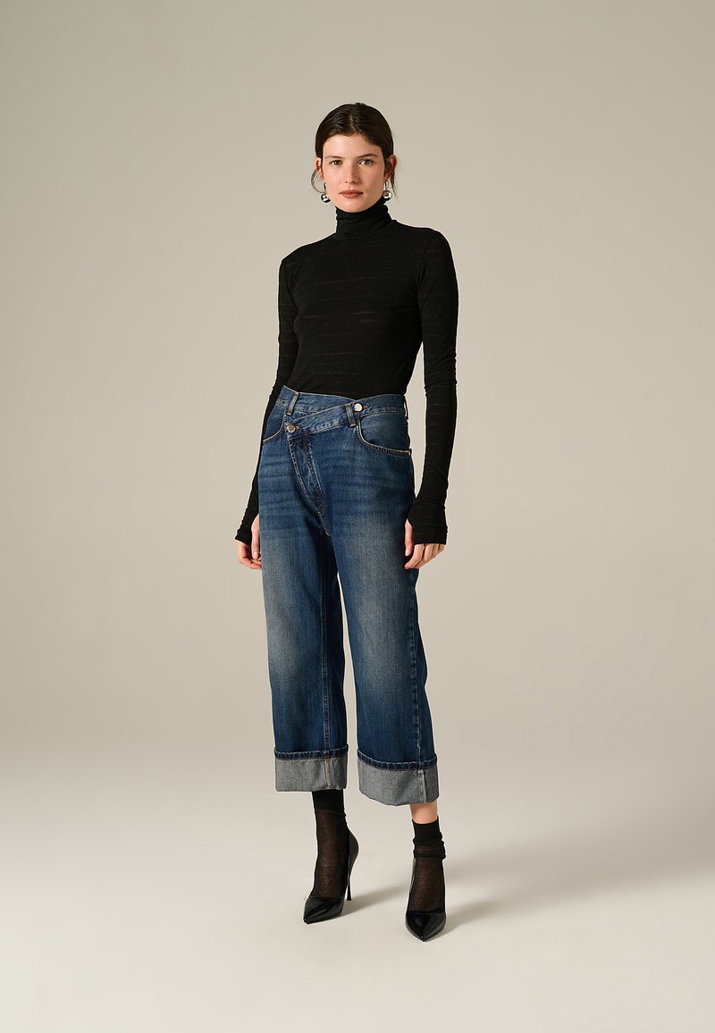 Donna in piedi che indossa un dolcevita nero, jeans a gamba larga blu con risvolto, calze nere trasparenti e décolleté neri a punta su uno sfondo beige.