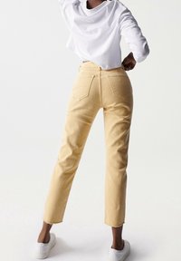 Jeans de algodón amarillos con corte recto, dobladillo deshilachado y bolsillos traseros. Combinados con una camiseta blanca de manga larga y zapatillas blancas.